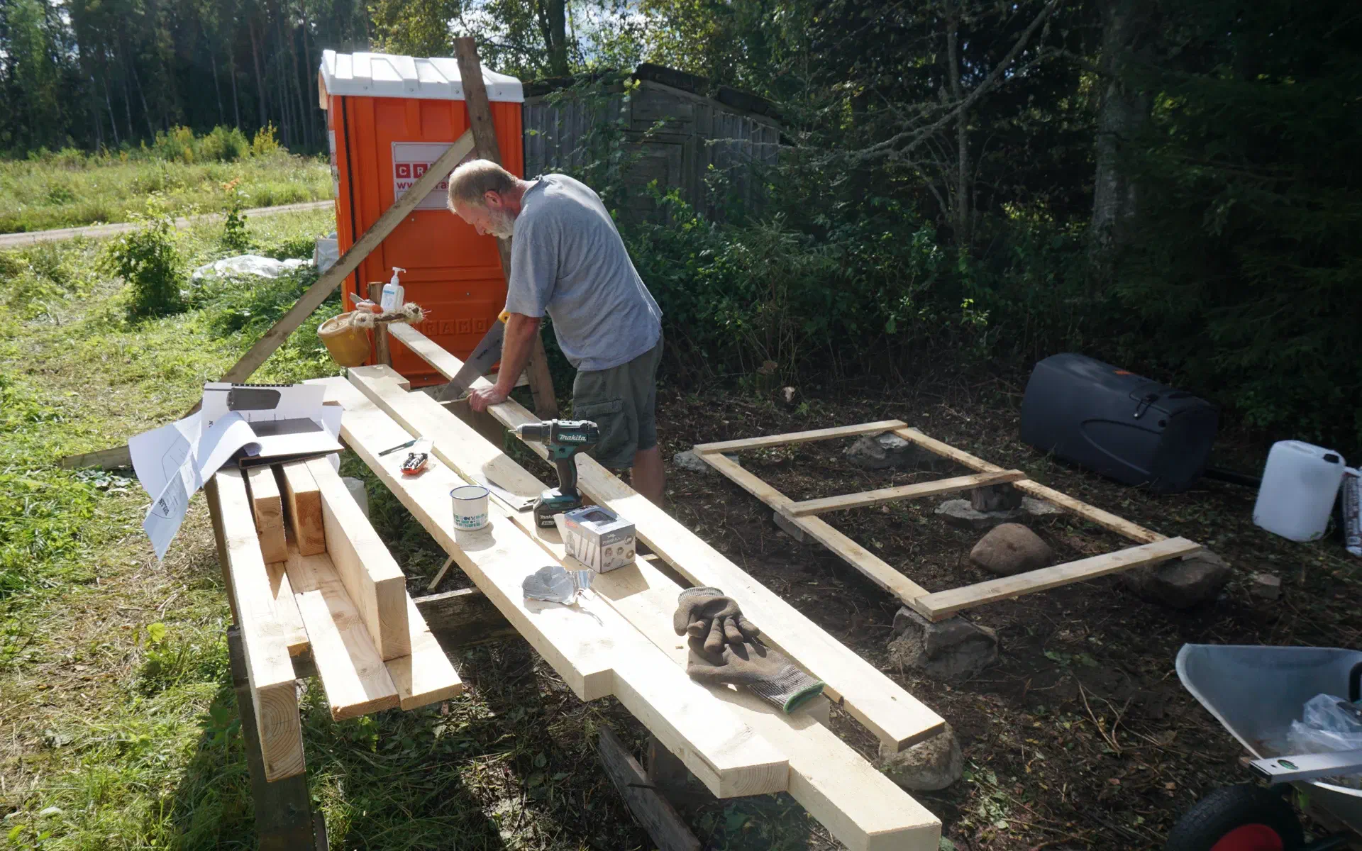 Tobias is sawing a board for the outhouse. In the background, the foundation is already complete.