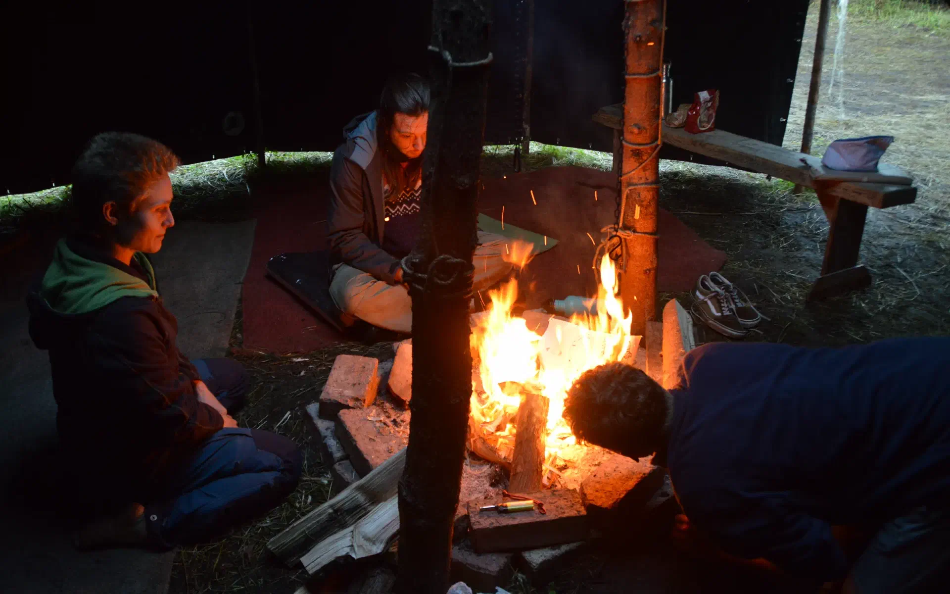 Zwei Menschen sitzen in einer Jurte am Feuer, eine dritte Person beugt sich vor, um das Feuer anzupusten.
