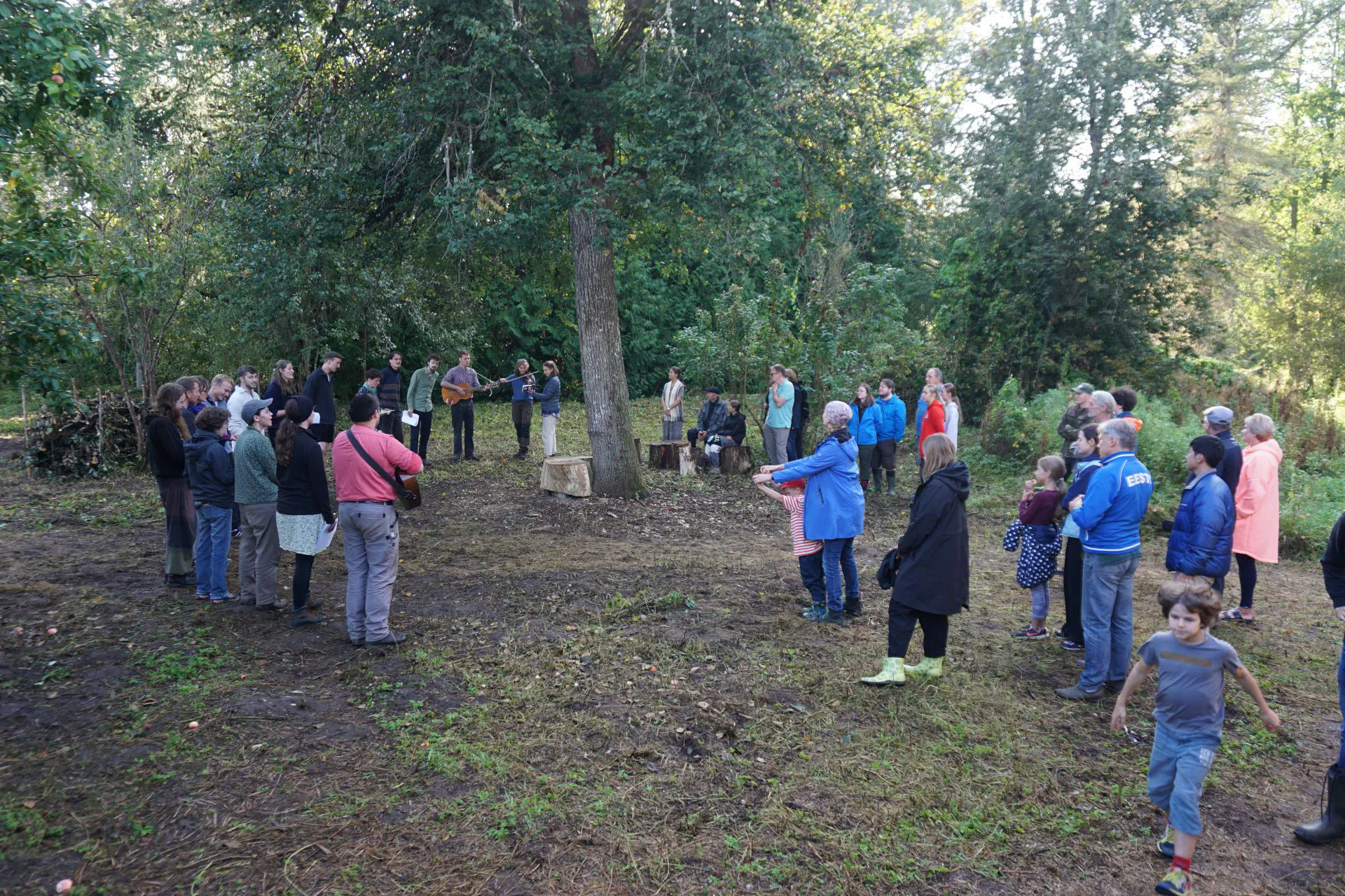 The farm team stands in a semi-circle around the lime tree, guitars and a violin can be seen. The guests watch, children play.