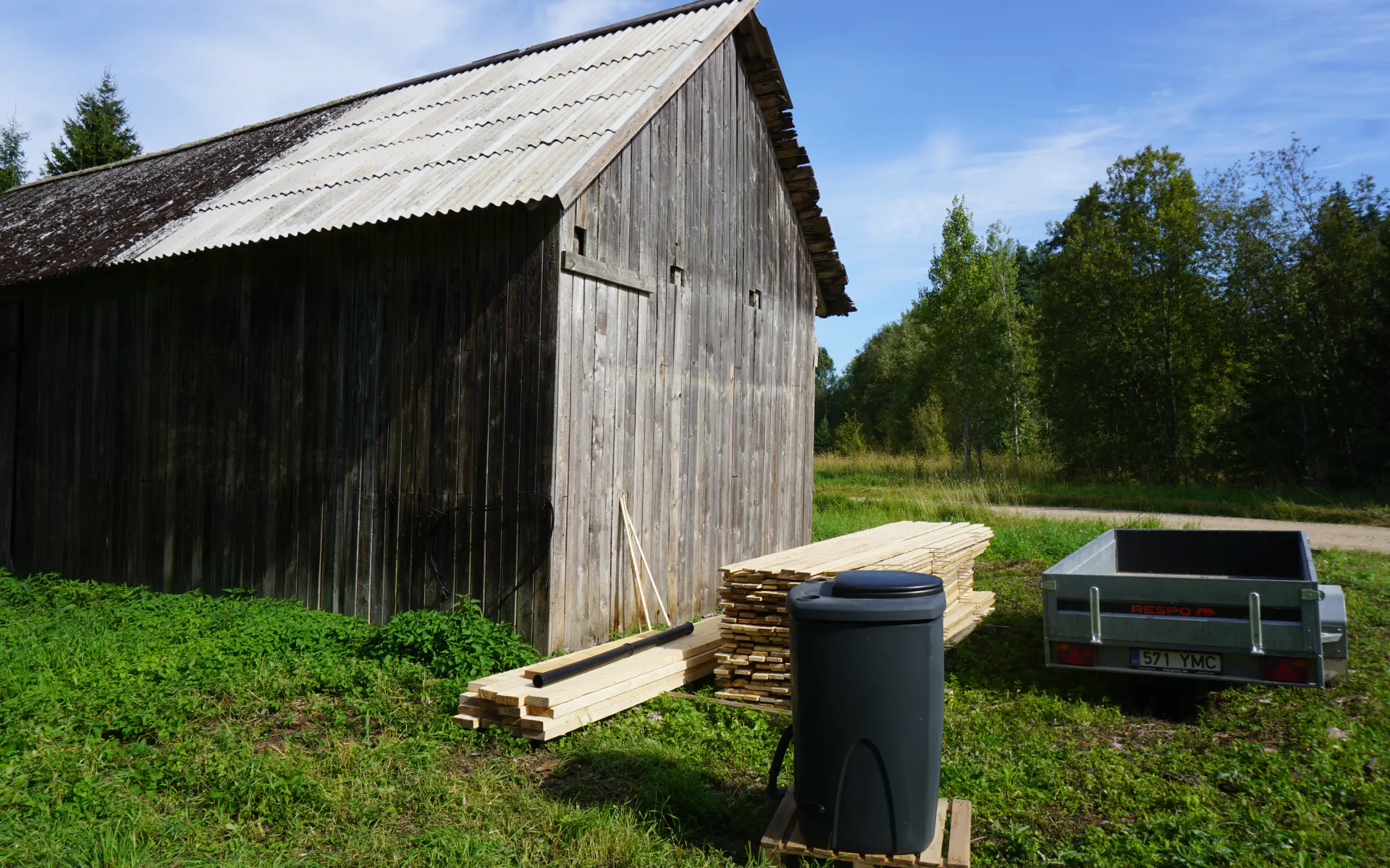 Neben der Scheune liegen Balken und Bretter für den Bau des Klohäuschens bereit. Im Vordergrund steht der Kompostklo-Behälter.