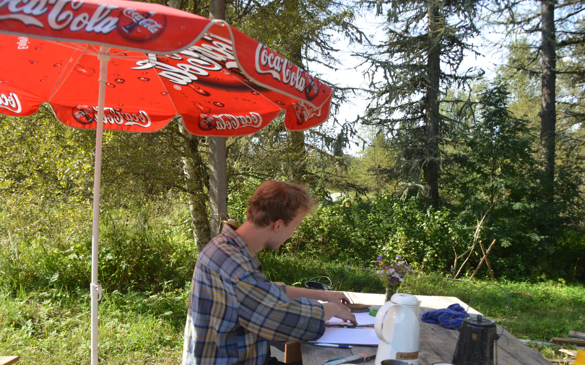 Klaas sitzt an einem Tisch mit Kaffee und Papier und zeichnet das Klohäuschen.