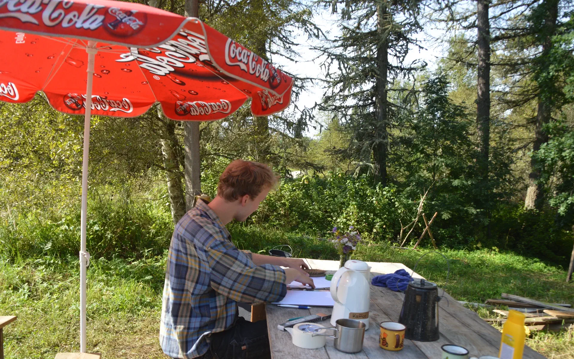 Klaas sits at a table with coffee and paper and draws the outhouse.