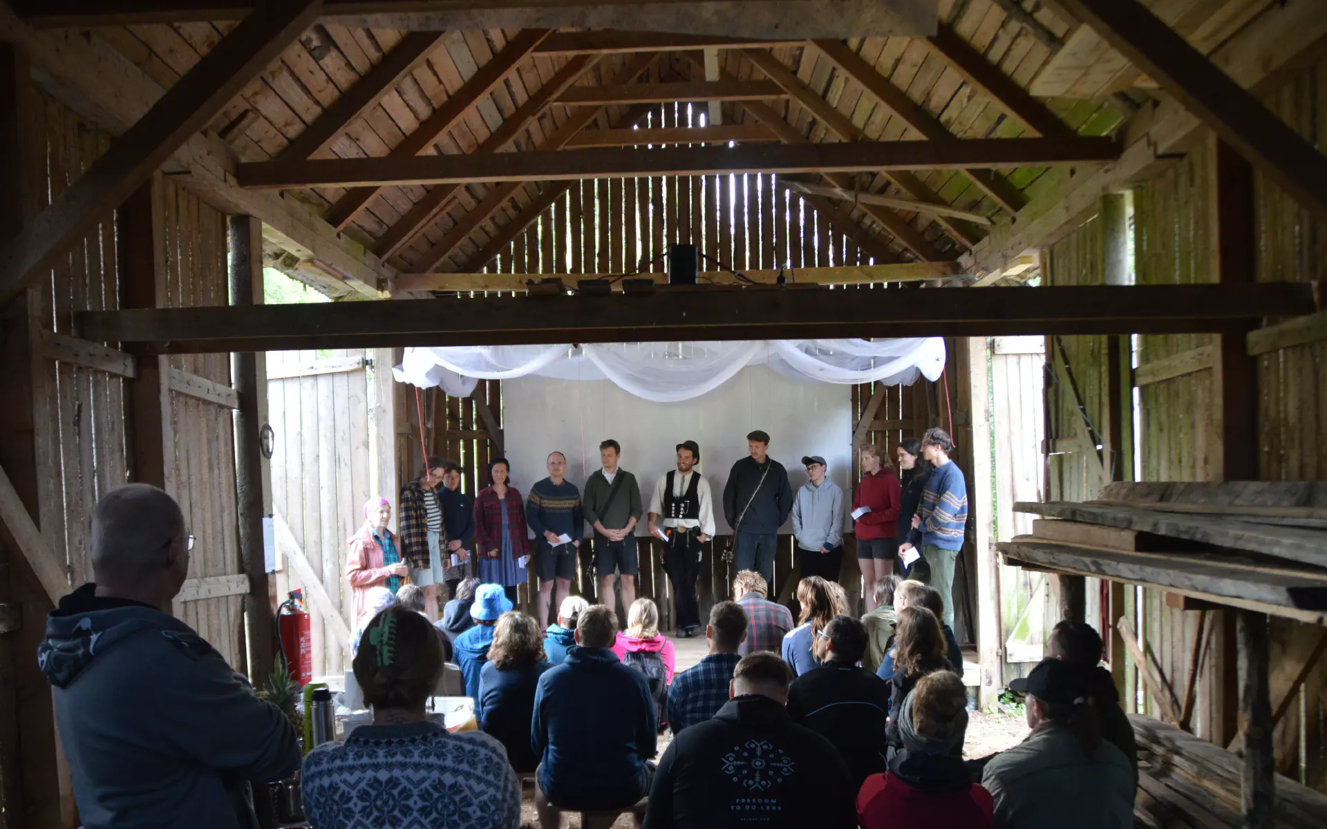 Auf der Bühne in der Scheune stehen elf Menschen, die etwas vortragen. Davor sitzt das Publikum auf Bänken.