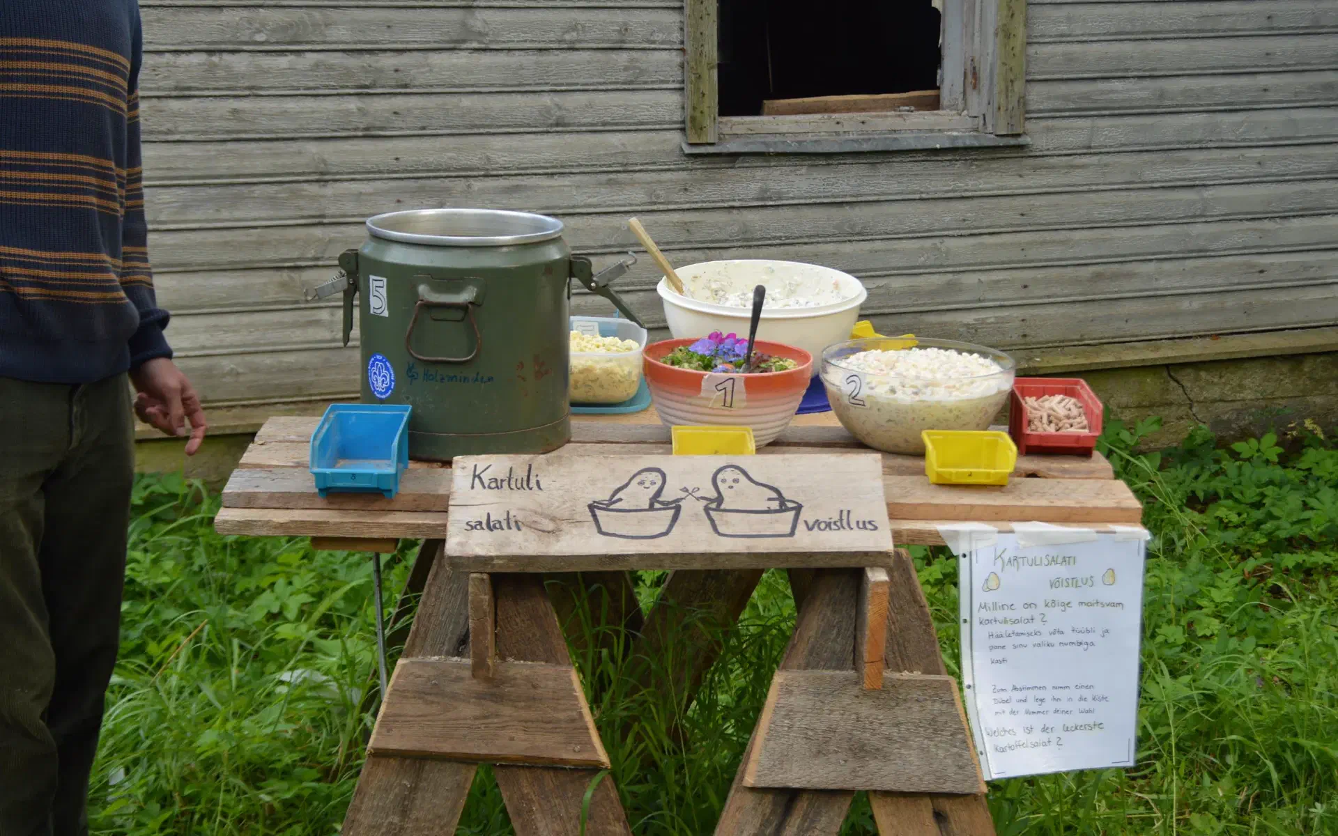 Several bowls of potato salad are on the buffet table. In the foreground is a painted sign with two potatoes fighting.