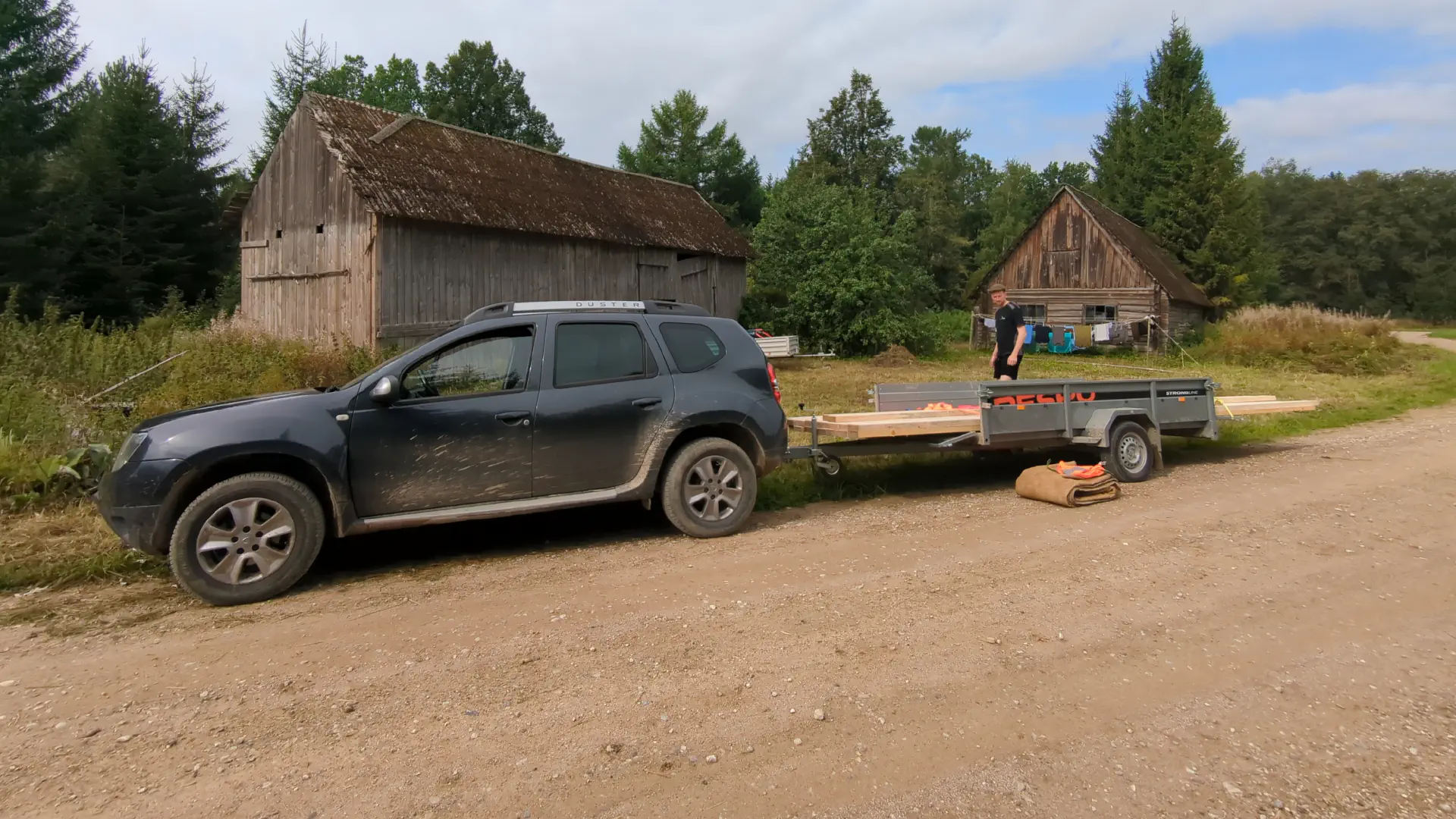 In front of the barn and the stable is a car with lots of wood on the trailer