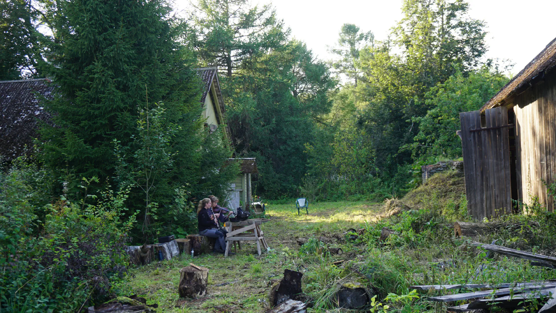 Two women sit in a mowed aisle between the main house and the barn and play the violin and clarinet