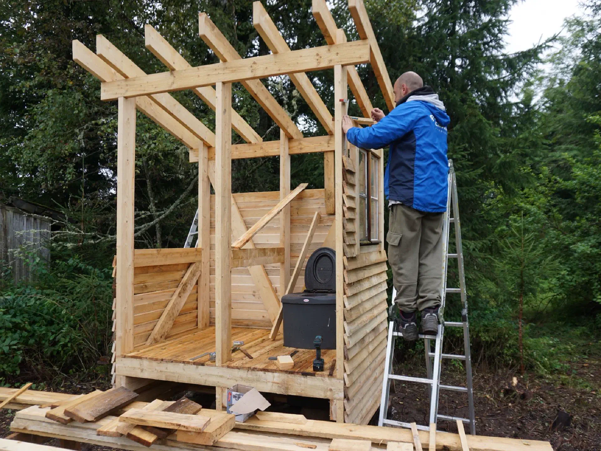 Jonas is standing on a ladder by the outhouse and taking measurements.