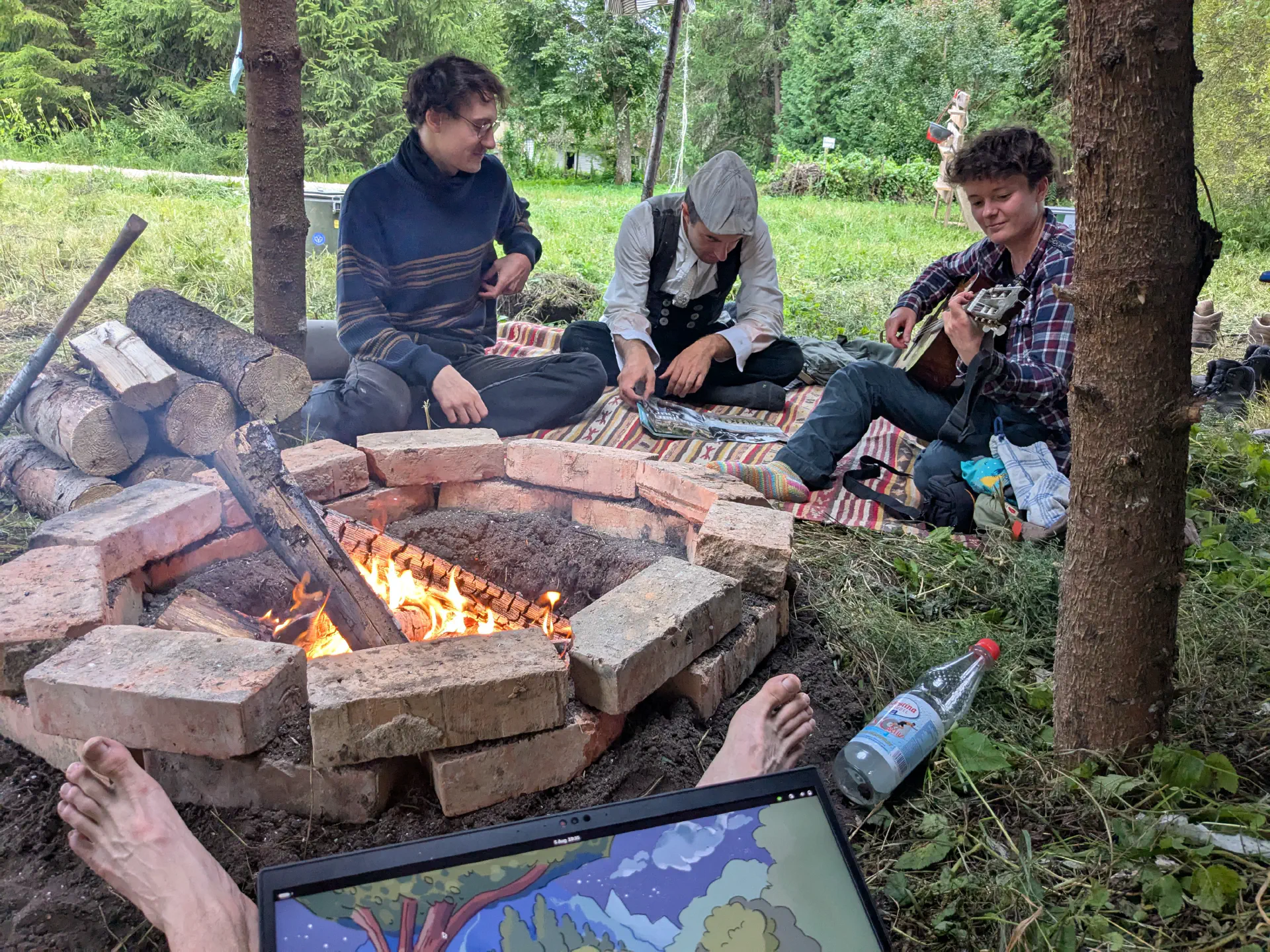 Behind a brick fireplace in a large black tent, three people are sitting reading or playing guitar.
