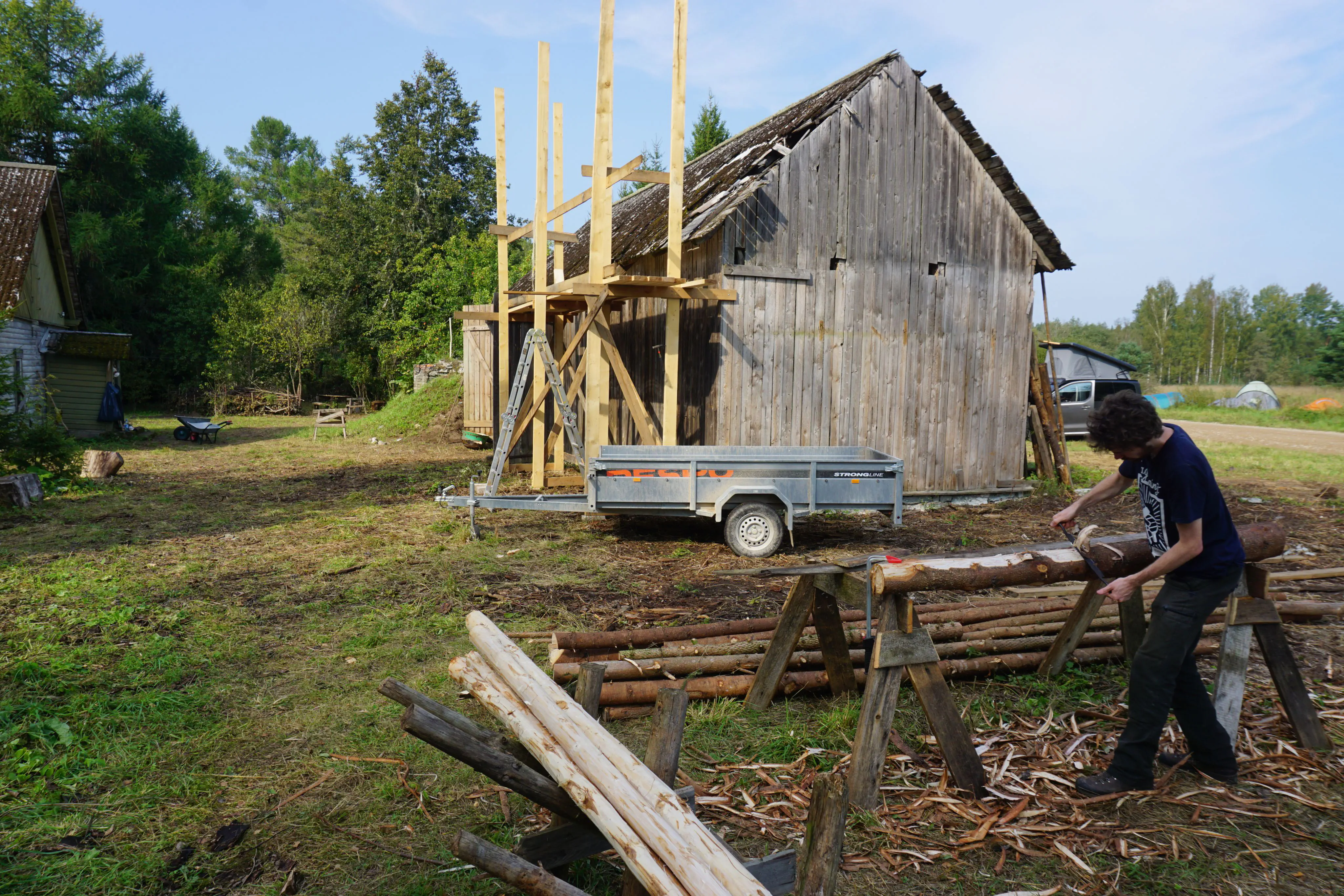 A man peeling a log in front of a farm house under construction